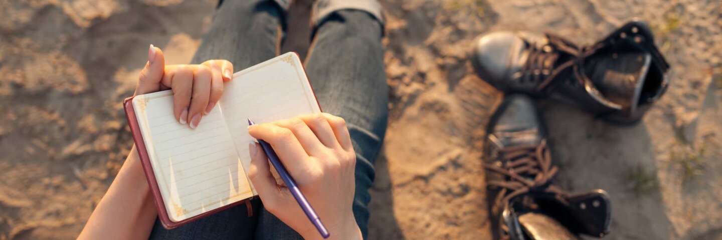 Woman writes in book while sitting in sand.