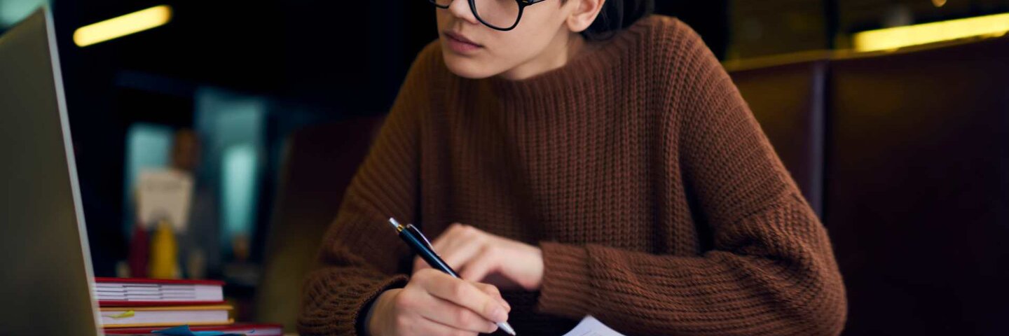 Female student in glasses taking notes while looking at laptop.