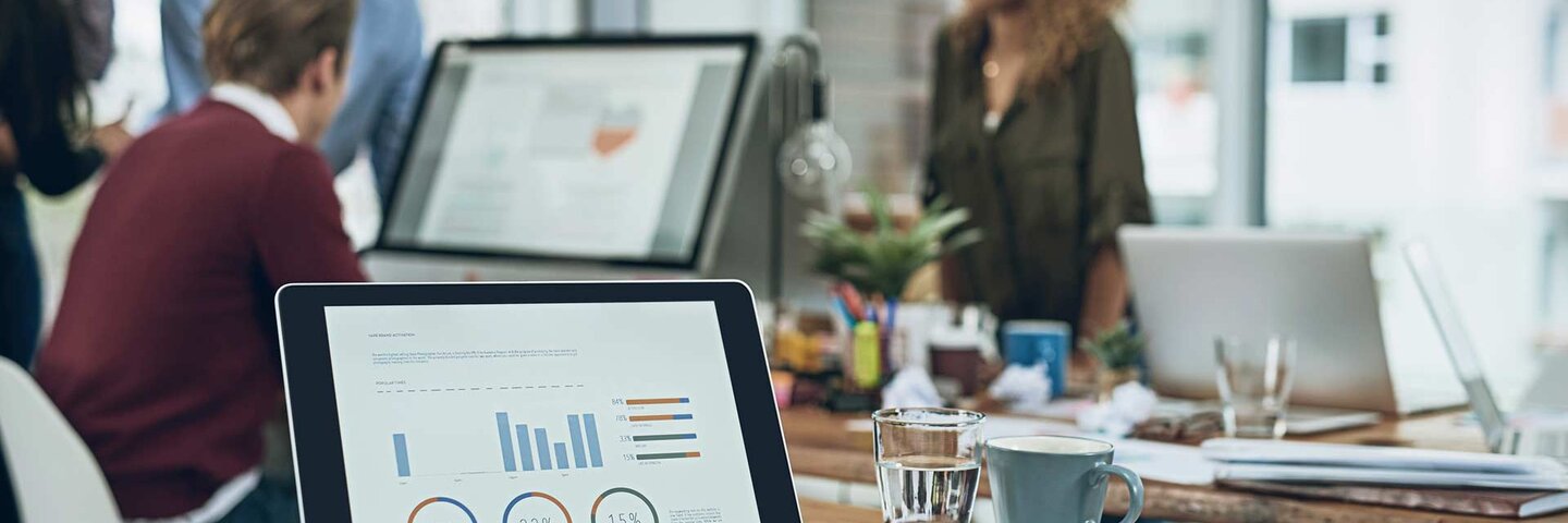 Closeup shot of a laptop with graphs on the screen and a team working in the background